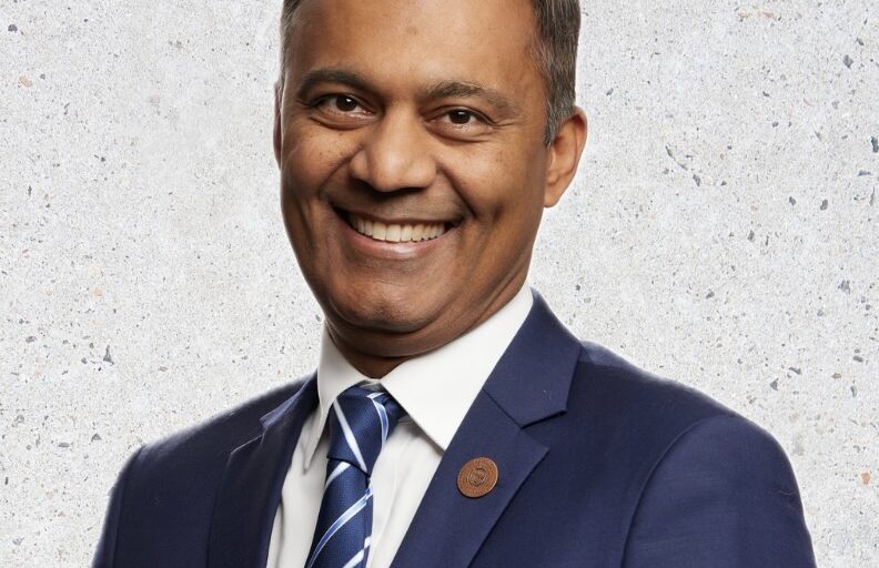 This is an image of Amish Shah, candidate for Arizona's 1st Congressional District. He is pictured in a blue suit standing in front of a white background.